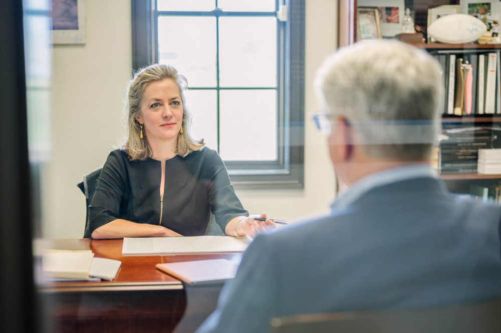 A woman with blonde hair, wearing a black top, sits at a desk in an office and looks at a man whose back is to the camera. A window is behind her and a bookshelf is to her right.
