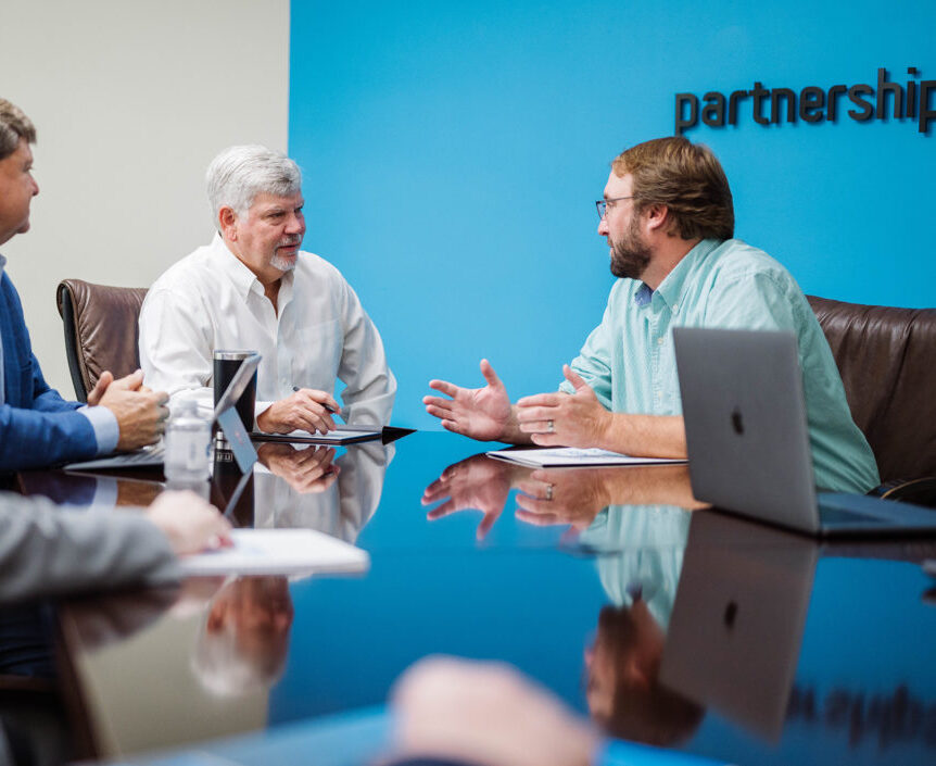 Three men sit around a highly reflective conference table during a meeting. The man on the right, with a beard and glasses, gestures with his hands as he speaks to the man in the center, who has gray hair and holds a pen. A bright blue wall behind them features the word "partnership" in black letters.