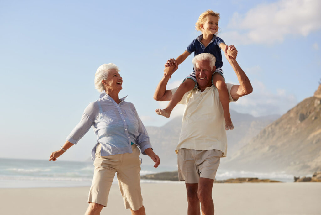 A joyful senior couple is on a sandy beach with their grandchild. The grandfather, wearing a polo shirt, smiles as he carries the young, blond-haired boy on his shoulders. The grandmother, in a striped shirt, walks beside them, smiling and looking up at the boy. The ocean is to their left and coastal hills are in the background.