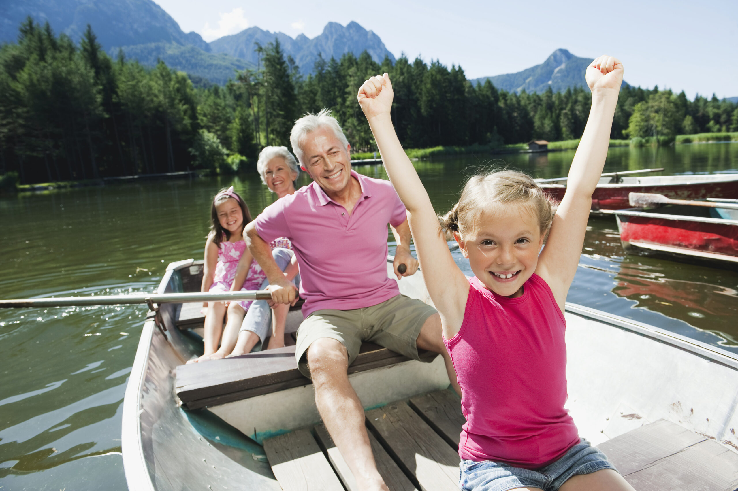 Two children and two older adults smile while rowing a boat on a calm lake surrounded by trees and mountains. The young girl in front raises her arms excitedly, and the others sit behind her enjoying the sunny day.